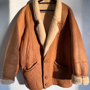 May include: A brown leather jacket with a cream-colored shearling lining and collar. The jacket has two front pockets, button closures, and shearling trim on the cuffs. It is hanging on a black hanger.