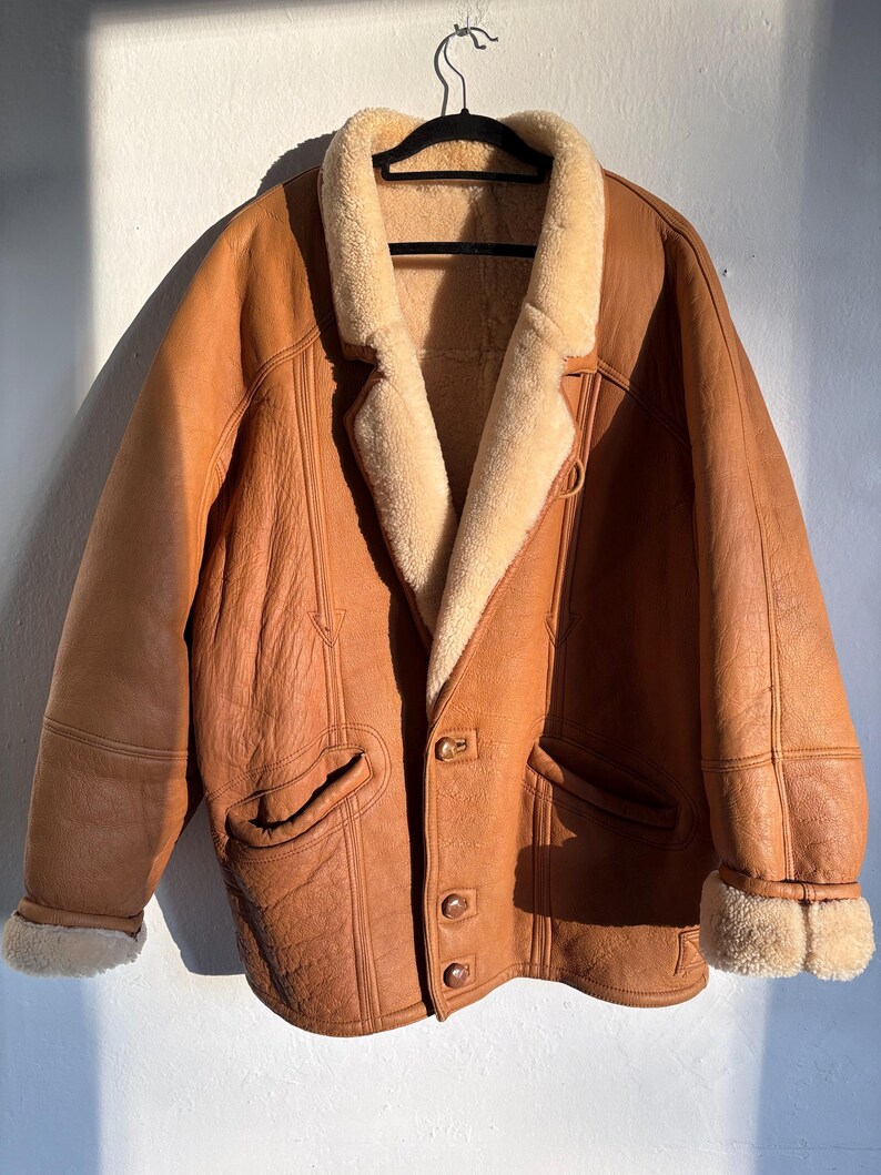 May include: A brown leather jacket with a cream-colored shearling lining and collar. The jacket has two front pockets, button closures, and shearling trim on the cuffs. It is hanging on a black hanger.