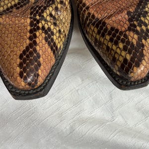 Puede incluir: Primer plano de las puntas de un par de botas de vaquero. Las botas presentan un estampado de piel de serpiente en tonos marrones, beige y amarillos. Las suelas son negras y el cuero tiene un aspecto texturizado.