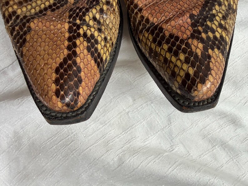 Puede incluir: Primer plano de las puntas de un par de botas de vaquero. Las botas presentan un estampado de piel de serpiente en tonos marrones, beige y amarillos. Las suelas son negras y el cuero tiene un aspecto texturizado.