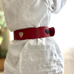 May include: A red leather belt with three heart-shaped accents in cream and navy blue. The belt is worn around the waist over a white linen shirt. The belt has a bow-shaped closure.