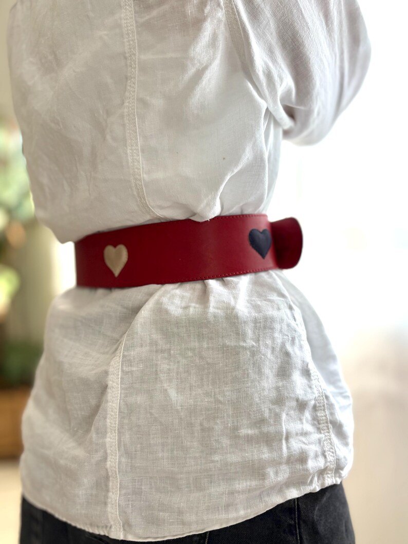 May include: A red leather belt with three heart-shaped accents in cream and navy blue. The belt is worn around the waist over a white linen shirt. The belt has a bow-shaped closure.
