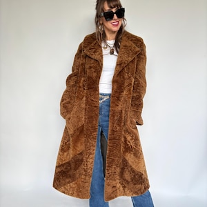 Könnte beinhalten: Ein langer, brauner Kunstfellmantel mit Reverskragen und strukturierter Optik. Der Mantel wird über einem weißen Oberteil und einer blauen Jeans getragen. Der Mantel hat eine knielange Länge und eine lockere Passform, geeignet für kühleres Wetter.