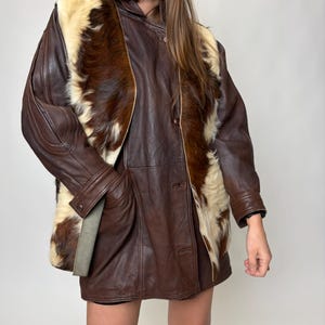 May include: A brown leather jacket with a fur-like panel on the front. The jacket has a button-up closure and long sleeves. The fur panel is a mix of brown and cream colors. The jacket is worn over a dark skirt and black boots.