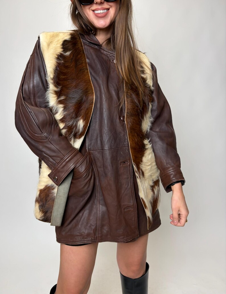 May include: A brown leather jacket with a fur-like panel on the front. The jacket has a button-up closure and long sleeves. The fur panel is a mix of brown and cream colors. The jacket is worn over a dark skirt and black boots.