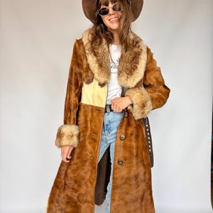 May include: A long brown coat with a textured fur-like finish, featuring a large tan faux fur collar and matching cuffs. The coat has a front button closure, a light-coloured panel on the chest, and is worn over a white t-shirt and blue jeans.