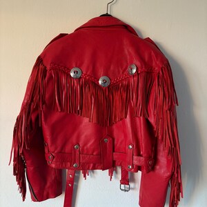 Op de afbeelding: Een rode leren jas met franjes en zilverkleurige knopen. De jas heeft een klassieke bikerstijl met een reverskraag, een riem in de taille en een ritssluiting. De achterkant van de jas heeft decoratief gevlochten leer.