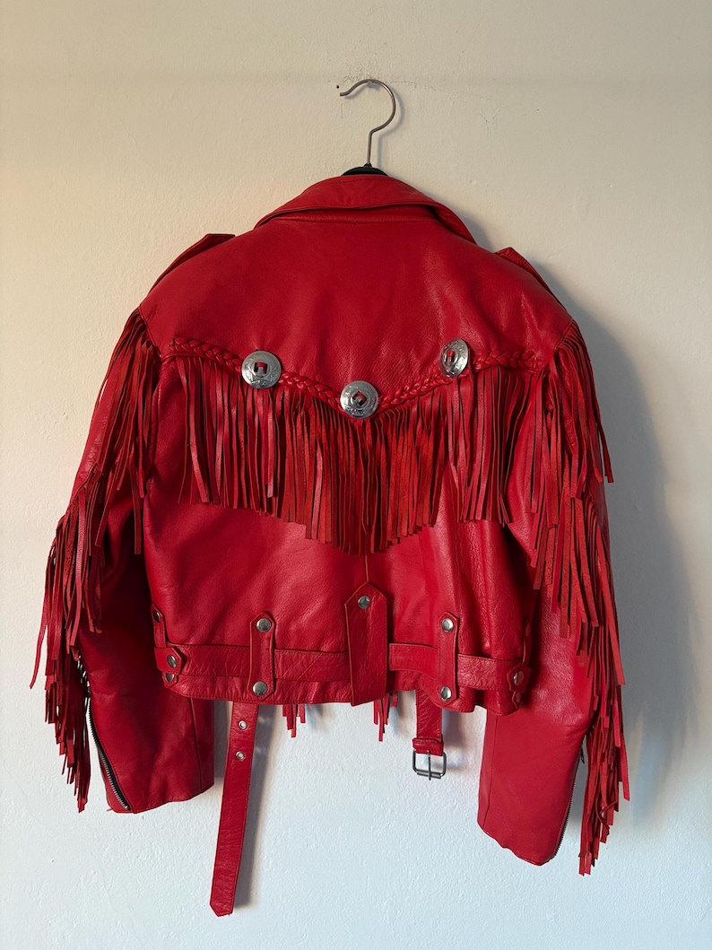 Op de afbeelding: Een rode leren jas met franjes en zilverkleurige knopen. De jas heeft een klassieke bikerstijl met een reverskraag, een riem in de taille en een ritssluiting. De achterkant van de jas heeft decoratief gevlochten leer.