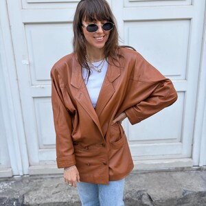 May include: A tan leather blazer featuring a double-breasted front with two buttons, wide lapels, and long sleeves with cuffs. The jacket is worn over a white top and light blue denim jeans, paired with oval-shaped sunglasses and layered necklaces.