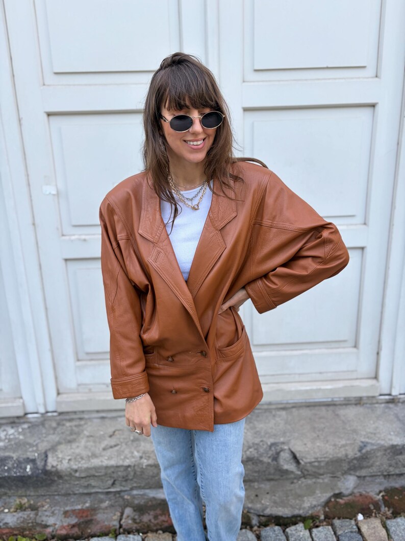 May include: A tan leather blazer featuring a double-breasted front with two buttons, wide lapels, and long sleeves with cuffs. The jacket is worn over a white top and light blue denim jeans, paired with oval-shaped sunglasses and layered necklaces.