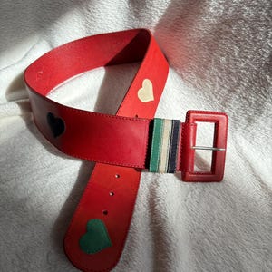 May include: A red leather belt with a square buckle. The belt features a black, white, and green striped accent and heart-shaped appliqu&eacute;s in black, white, and green. The belt is approximately 1.5 inches wide.