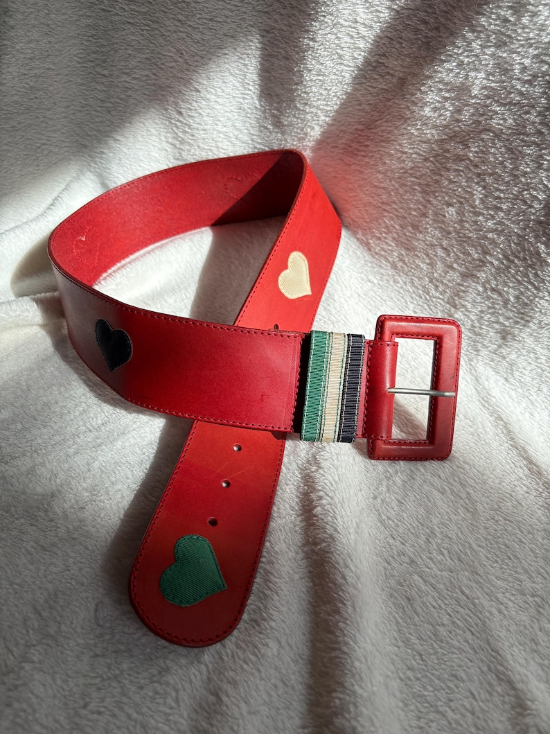 May include: A red leather belt with a square buckle. The belt features a black, white, and green striped accent and heart-shaped appliqu&eacute;s in black, white, and green. The belt is approximately 1.5 inches wide.