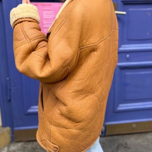 May include: A tan leather jacket with a cream-colored shearling lining on the collar and cuffs. The jacket has a classic design with a button closure and a warm, inviting appearance. The jacket is worn over light blue jeans.