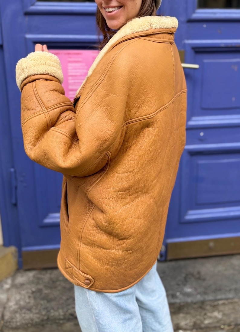 May include: A tan leather jacket with a cream-colored shearling lining on the collar and cuffs. The jacket has a classic design with a button closure and a warm, inviting appearance. The jacket is worn over light blue jeans.