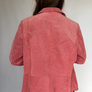 May include: A dusty rose-colored suede blazer with a classic collar and long sleeves. The back view of the jacket shows the tailored seams and a slightly textured finish. The blazer is part of a vintage clothing collection.