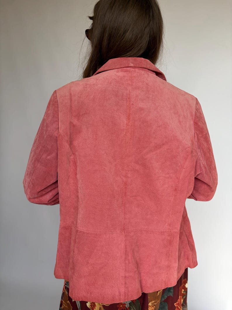 May include: A dusty rose-colored suede blazer with a classic collar and long sleeves. The back view of the jacket shows the tailored seams and a slightly textured finish. The blazer is part of a vintage clothing collection.