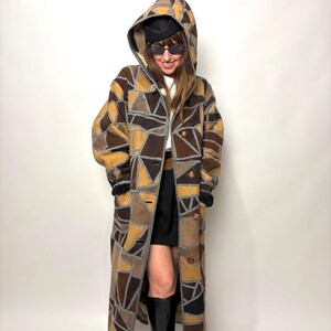 May include: A long patchwork coat with a hood. The coat features a geometric pattern in shades of brown, tan, and gray. It has a button-up front and a cozy, hooded design. The coat is knee-length and appears to be made of a soft, textured material.