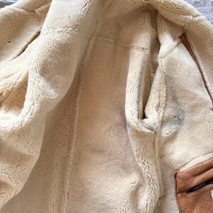 May include: A tan shearling jacket with a cream-colored, fluffy interior. The jacket features a brown leather pocket with a black zipper. The interior is visible, showcasing the soft, warm texture of the material.
