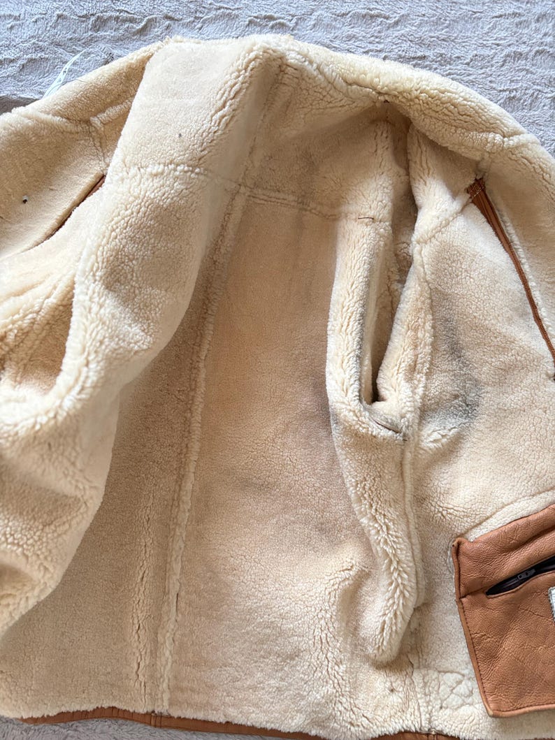 May include: A tan shearling jacket with a cream-colored, fluffy interior. The jacket features a brown leather pocket with a black zipper. The interior is visible, showcasing the soft, warm texture of the material.