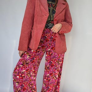 May include: A dusty rose blazer with a two-button closure and front pockets. Paired with wide-leg pants featuring a vibrant floral pattern in shades of pink, red, and yellow. The outfit includes a black graphic t-shirt.