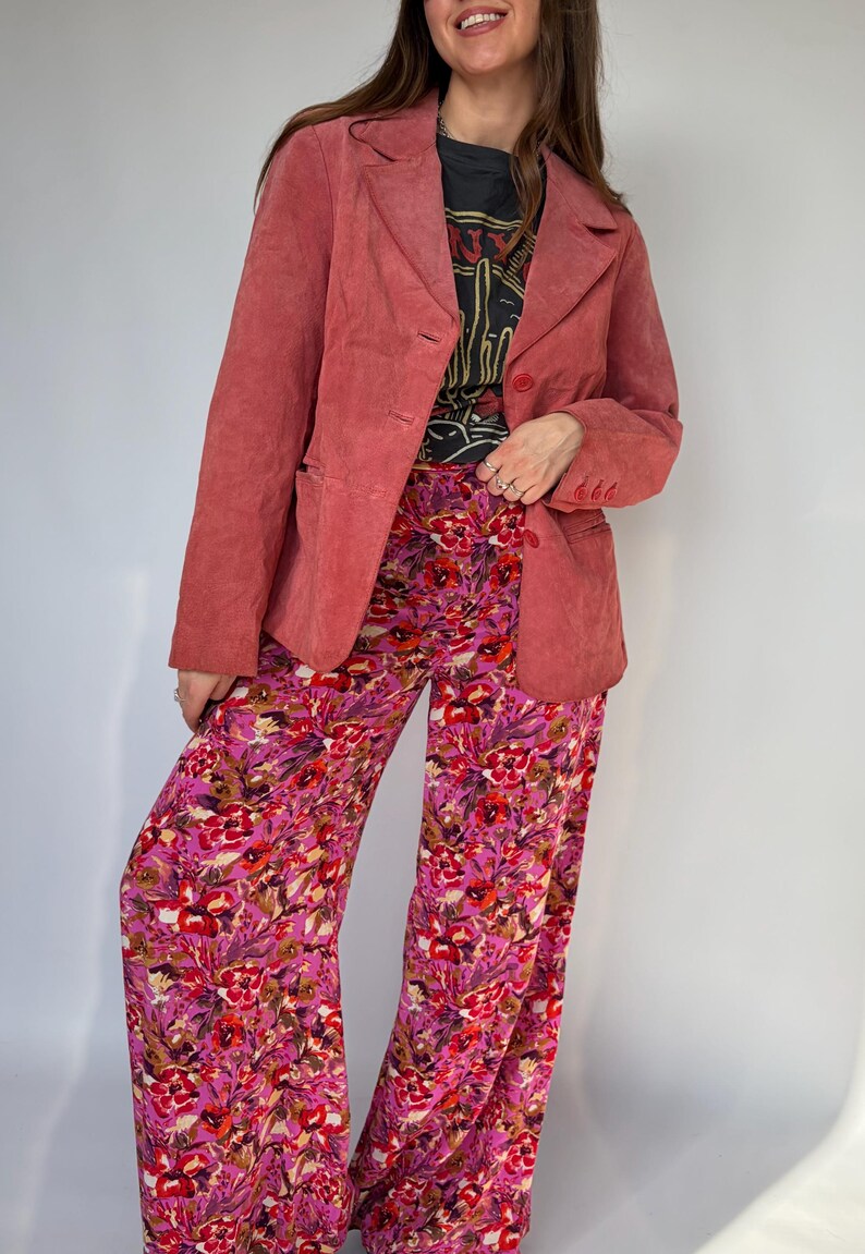 May include: A dusty rose blazer with a two-button closure and front pockets. Paired with wide-leg pants featuring a vibrant floral pattern in shades of pink, red, and yellow. The outfit includes a black graphic t-shirt.