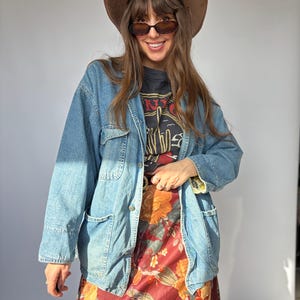 May include: A light blue denim jacket with two chest pockets and a button-up front. The jacket is worn over a black graphic tee and a long floral print dress. A brown hat and sunglasses complete the look.