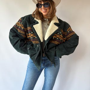 Könnte beinhalten: Eine dunkelgrüne Wildlederjacke mit cremefarbenem Sherpa-Kragen und einem horizontalen Brustband mit geometrischem Muster in Braun, Schwarz und Kastanienbraun. Die Jacke wird mit einer blauen Jeans, einem cremefarbenen Cowboyhut und einer Sonnenbrille kombiniert.