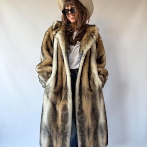 May include: A long, faux fur coat featuring a multi-tonal pattern of brown, tan, gray, and white vertical stripes. The coat has a wide collar, long sleeves, and an open front design. It is worn over a white t-shirt and blue denim jeans.
