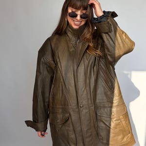 K&ouml;nnte beinhalten: Braune Lederjacke mit hohem Kragen, Rei&szlig;verschluss und mehreren Taschen. Die Jacke hat eine lockere Passform und eine Vintage-&Auml;sthetik. Die &Auml;rmel haben gekn&ouml;pfte Manschetten. Die Jacke wird von einer Person mit Sonnenbrille getragen.