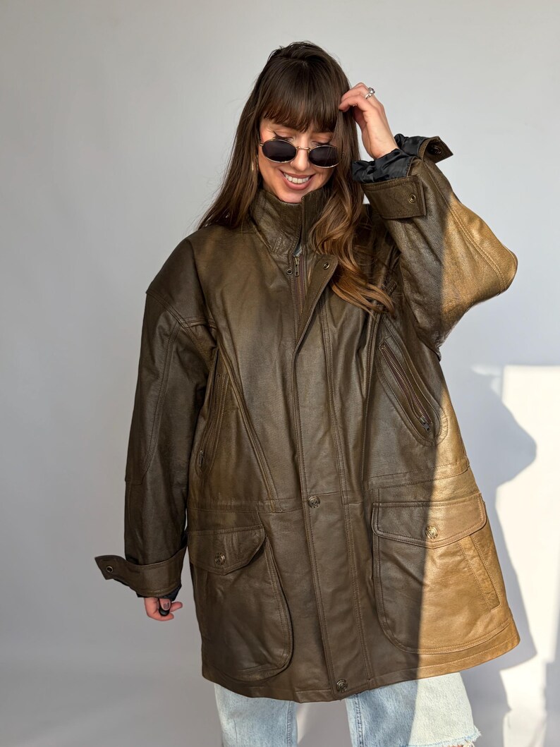 K&ouml;nnte beinhalten: Braune Lederjacke mit hohem Kragen, Rei&szlig;verschluss und mehreren Taschen. Die Jacke hat eine lockere Passform und eine Vintage-&Auml;sthetik. Die &Auml;rmel haben gekn&ouml;pfte Manschetten. Die Jacke wird von einer Person mit Sonnenbrille getragen.