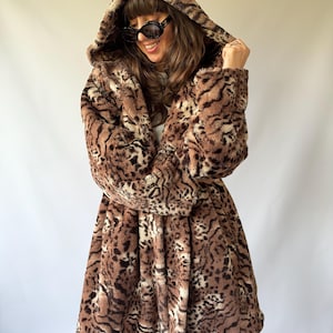 May include: A hooded jacket made of faux fur featuring a brown, black, and cream animal print pattern. The jacket has long sleeves, a full hood, and a mid-thigh length, showcasing a dense, textured pile throughout the entire garment.