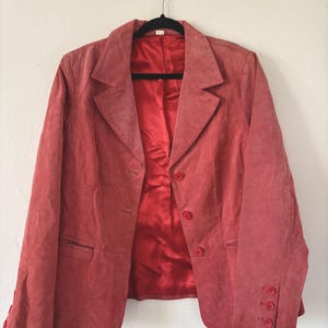 May include: A coral-colored blazer with a classic notched lapel and three-button closure. The blazer features a matching lining and buttons, and is hanging on a black hanger against a white wall.