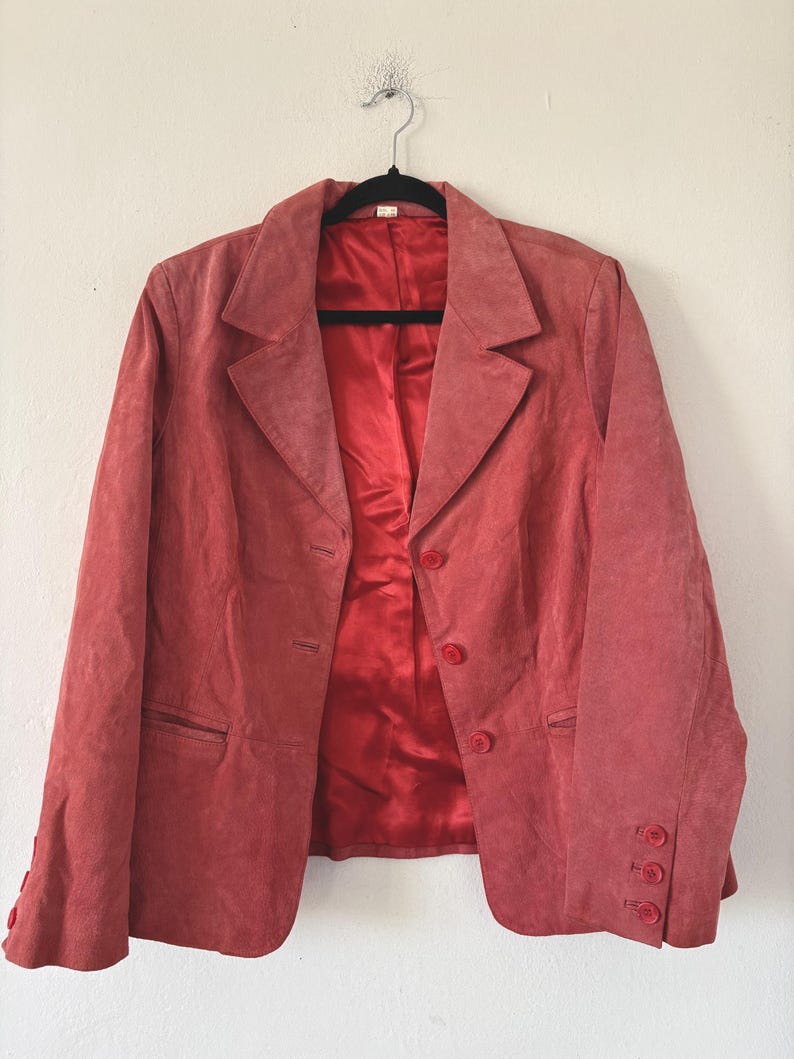 May include: A coral-colored blazer with a classic notched lapel and three-button closure. The blazer features a matching lining and buttons, and is hanging on a black hanger against a white wall.