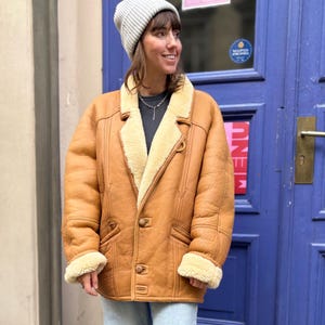 May include: A tan shearling coat with a light beige interior and trim. The coat has a button closure, two front pockets, and a notched collar. The person is wearing a gray beanie and light blue jeans.
