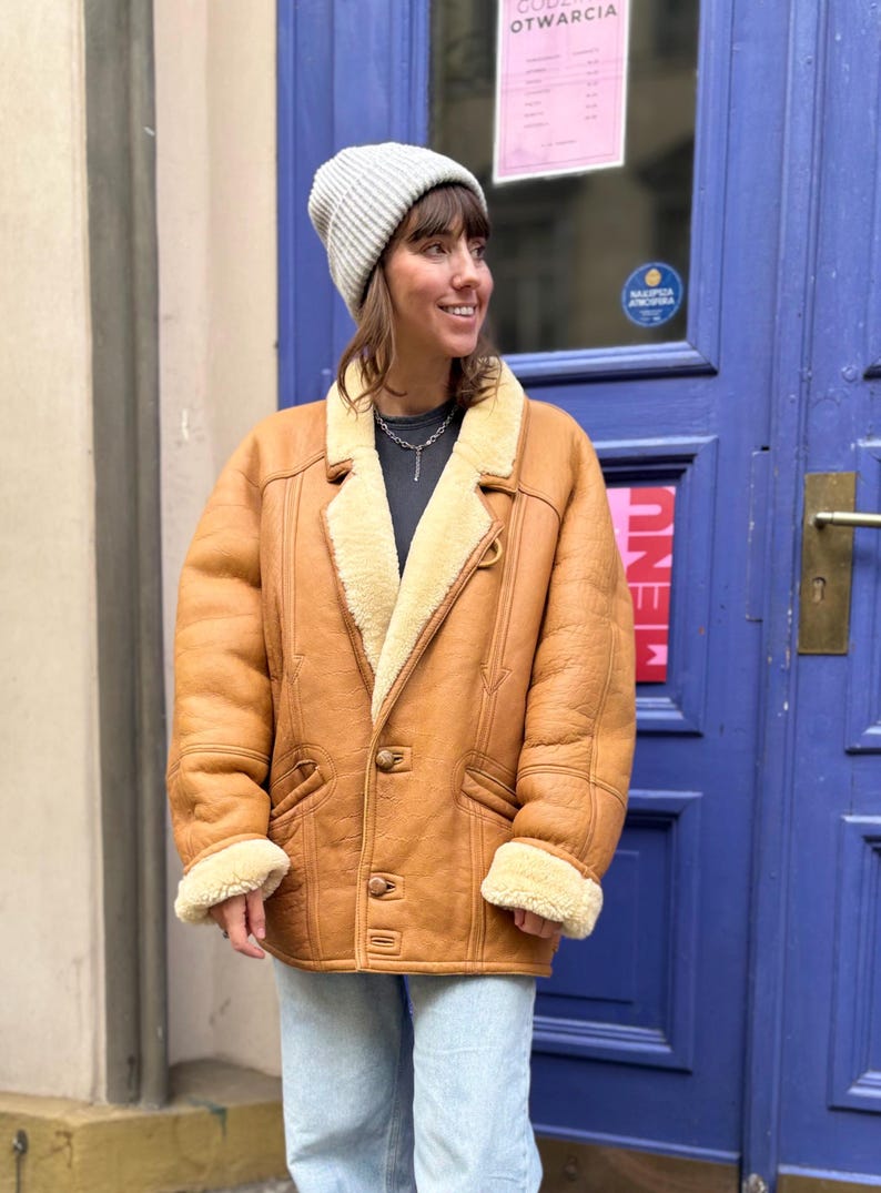 May include: A tan shearling coat with a light beige interior and trim. The coat has a button closure, two front pockets, and a notched collar. The person is wearing a gray beanie and light blue jeans.
