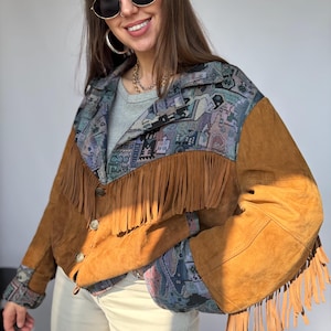 May include: A brown suede jacket with fringe detailing. The jacket features a patterned fabric on the shoulders and cuffs, with a button-up front. The fringe hangs from the chest and sleeves. The jacket is a medium brown color.