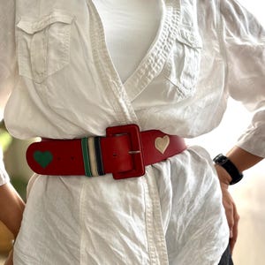 May include: A red leather belt with a large square buckle. The belt features green and cream heart-shaped accents, along with a striped section of green, navy, and cream. The belt is worn over a white shirt.