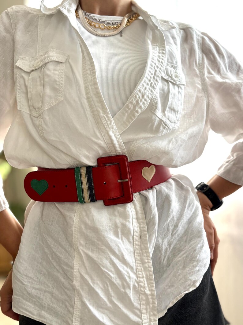 May include: A red leather belt with a large square buckle. The belt features green and cream heart-shaped accents, along with a striped section of green, navy, and cream. The belt is worn over a white shirt.