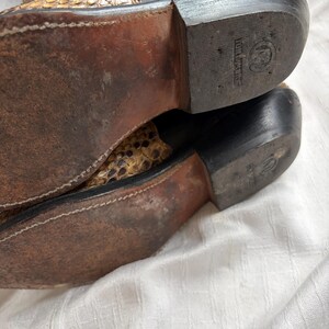 Puede incluir: Primer plano de las suelas de un par de botas de vaquero. Las botas son negras y marrones con un estampado de piel de serpiente en la parte superior. Las suelas est&aacute;n desgastadas y muestran signos de uso.