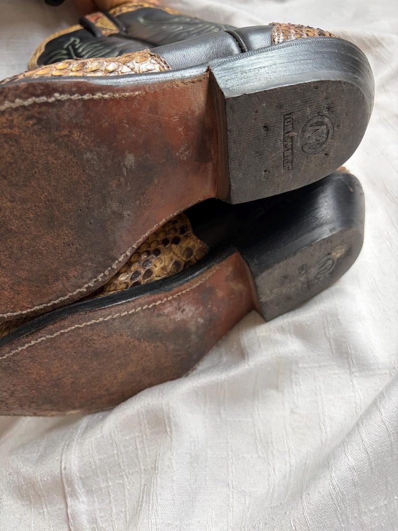 Puede incluir: Primer plano de las suelas de un par de botas de vaquero. Las botas son negras y marrones con un estampado de piel de serpiente en la parte superior. Las suelas est&aacute;n desgastadas y muestran signos de uso.