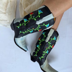 May include: A pair of black leather cowboy boots featuring a pointed toe and a stacked heel. The boots are decorated with vibrant green, blue, and red floral embroidery that climbs up the shaft. The lower foot area is a faded light blue color.