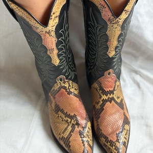 Puede incluir: Botas de vaquero negras con estampado de piel de serpiente en la puntera y la ca&ntilde;a. Las botas tienen punta afilada y costuras decorativas. El estampado de serpiente presenta tonos marrones, naranjas y amarillos.