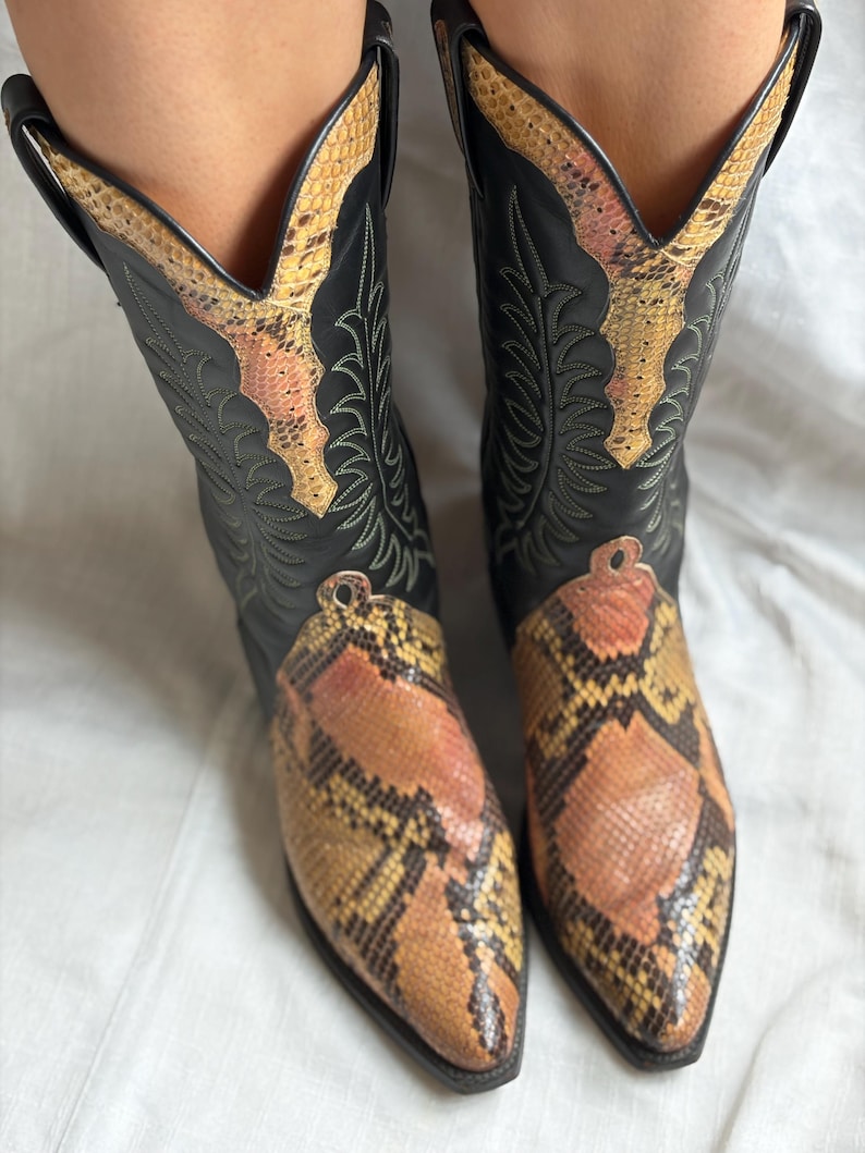 Puede incluir: Botas de vaquero negras con estampado de piel de serpiente en la puntera y la ca&ntilde;a. Las botas tienen punta afilada y costuras decorativas. El estampado de serpiente presenta tonos marrones, naranjas y amarillos.