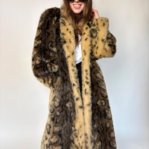 May include: A long faux fur coat with a leopard print design in shades of brown and tan. The coat has a light tan trim and a plush texture. The model is wearing a red beret and sunglasses.