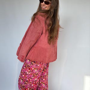 May include: A dusty rose suede blazer with button details is paired with vibrant floral print pants. The pants feature a mix of pink, red, and brown hues. The outfit is completed with dark sunglasses.