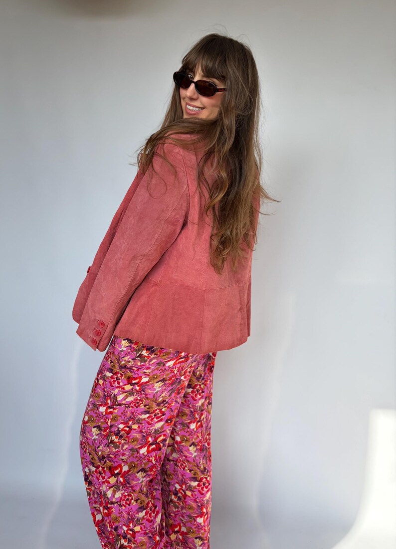 May include: A dusty rose suede blazer with button details is paired with vibrant floral print pants. The pants feature a mix of pink, red, and brown hues. The outfit is completed with dark sunglasses.