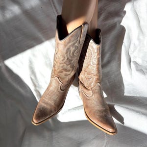 Può includere: Un paio di stivali da cowboy marrone chiaro con intricati disegni cuciti. Gli stivali hanno punte a punta e un piccolo tacco. La pelle sembra usurata, conferendo loro un aspetto vintage. Gli stivali sono su una superficie bianca.