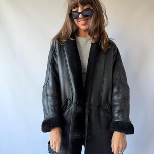 May include: A black leather coat featuring a thick black faux fur lining, a wide collar, and long sleeves with folded cuffs. The coat has a button-front closure and two side pockets. The exterior is made of black leather with visible seams.