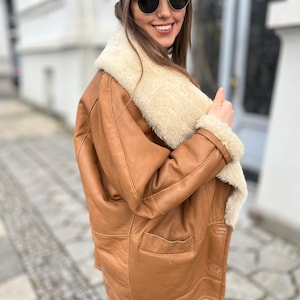 May include: A brown leather jacket with a white shearling collar and lining. The jacket has a zipper closure and two pockets. The person is wearing a brown baseball cap and sunglasses.
