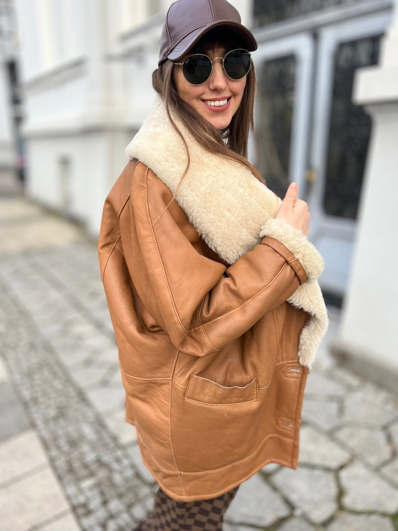May include: A brown leather jacket with a white shearling collar and lining. The jacket has a zipper closure and two pockets. The person is wearing a brown baseball cap and sunglasses.